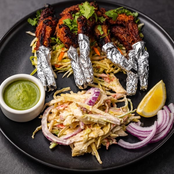 Chicken lollipops with cilantro, coleslaw, green sauce, lemon, and red onion on a black plate.