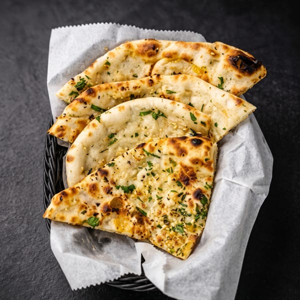 Slices of golden naan bread with herbs and sesame seeds in a basket on a dark surface.