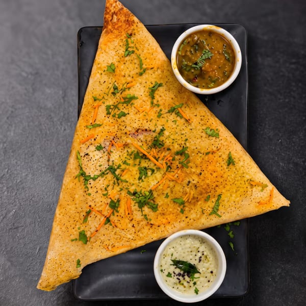 Triangular dosa garnished with cilantro and carrots, served with sambar and coconut chutney on a black plate.